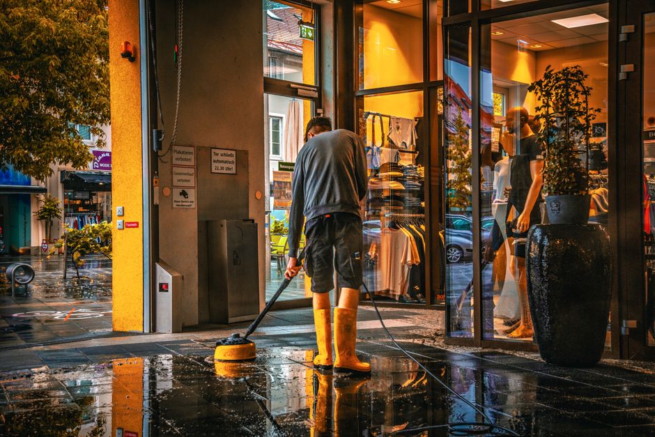 A person wearing yellow rubber boots and a gray sweatshirt is performing surface cleaning outside the entrance of a retail store in Barnet during daytime. They are using a pressure washer or similar c