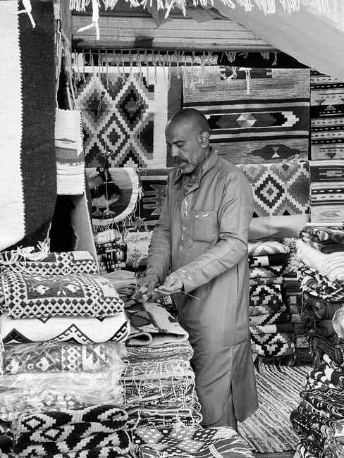 A man wearing a grey vest and trousers is inside a small shop or storage space, bent over while organizing or inspecting a stack of folded carpets and rugs. Surrounding him are various hanging and sta