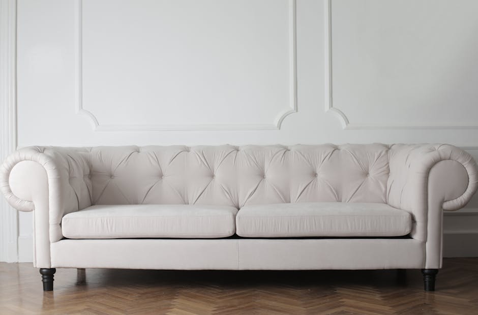 A white upholstered sofa with tufted backrest and rolled arms, positioned in front of a white paneled wall, on a wooden floor. The sofa appears clean and well-maintained, indicative of professional su